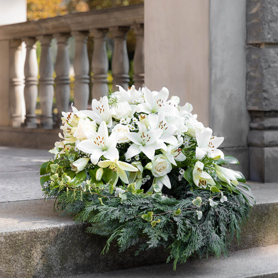 Weißer Trauerkranz mit Lilien und Rosen, arrangiert auf einer Steintreppe vor einem historischen Gebäude – symbolischer Abschiedsgruß bei einer Trauerfeier.
