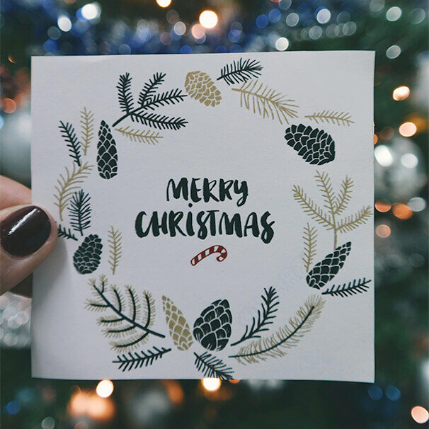 Weihnachtskarte mit der Aufschrift „Merry Christmas“ in schwarzer Schrift, umrahmt von einem Kranz aus Tannenzweigen und Zapfen, vor unscharfem, festlich beleuchtetem Hintergrund.