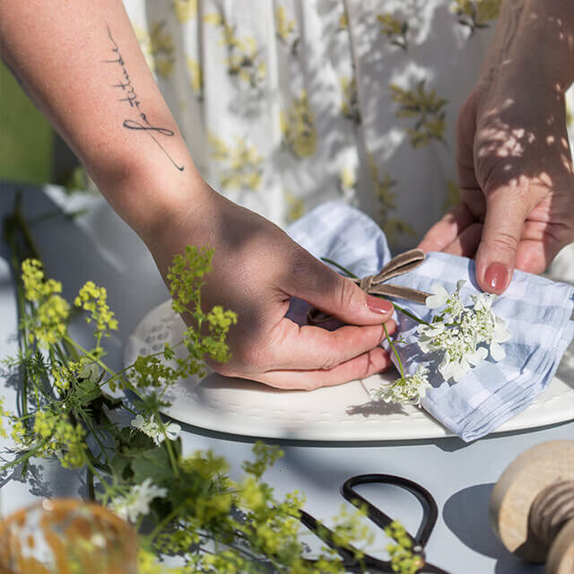 Tischdeko für Gartenparty - Step 6 Nahaufnahme von Händen, die eine karierte Stoffserviette mit einem kleinen Blumenstiel und einem Band verzieren.