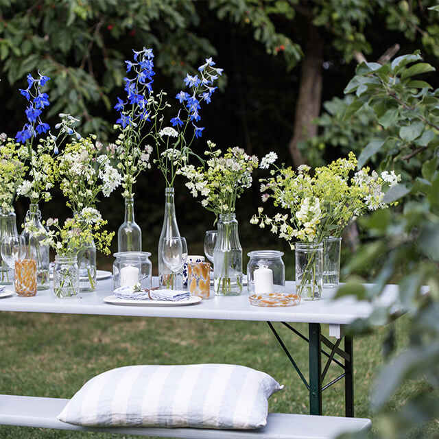 Tischdeko für Gartenparty Eine komplette Garten-Tafel im Boho-Stil mit zahlreichen Flaschenvasen voller Wildblumen, Kerzen und bunten Gläsern, darunter gestreifte Kissen auf der Bank.