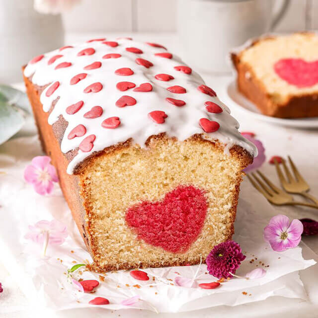 Fertiger Herzkuchen von Fleurop Aufgeschnittener Kuchen mit Herzüberraschung im Inneren, dekoriert mit Zuckerguss und Zuckerherzen, daneben Blüten und Besteck.