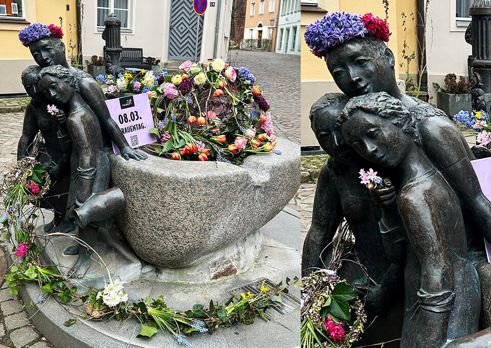 Figuren des Mägdebrunnens in Stralsund, verziert mit bunten Blumenarrangements und Kränzen anlässlich des Frauentags.