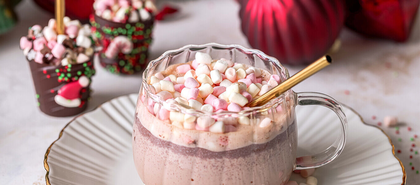 Ein weihnachtliches Arrangement mit einer heißen Schokolade in einer durchsichtigen Tasse, dekoriert mit bunten Mini-Marshmallows, vor einer festlichen Kulisse mit roten Weihnachtskugeln.