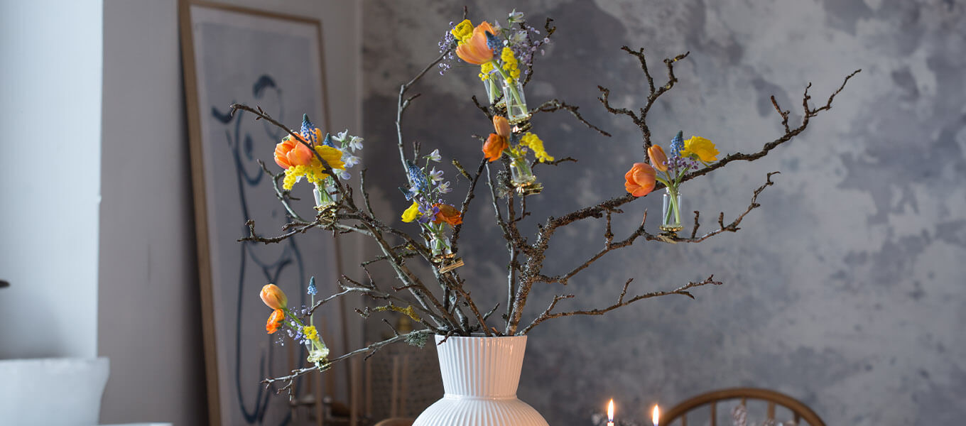 Weiße Vase gefüllt mit Ästen, an denen mithilfe kleiner Mini-Vasen Frühlingsblumen befestigt sind