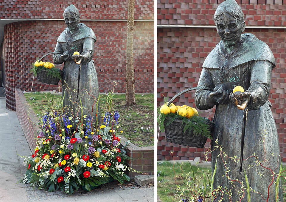 Zitronenjette-Statue in Hamburg mit Blumen zum Frauentag Statue der Zitronenjette in Hamburg, umgeben von bunten Frühlingsblumen und einem Korb voller Zitronen, geschmückt zum Frauentag.