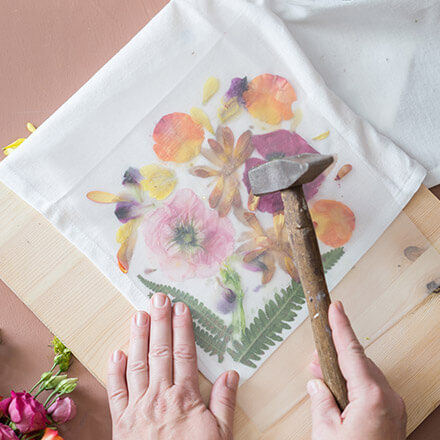 Zu sehen ist eine Hand, die mithilfe eines Hammers auf eine Stoffserviette hämmert . Unter der Stoffserviette liegen einzelne Blütenblätter, die in einem Muster angeordnet sind.