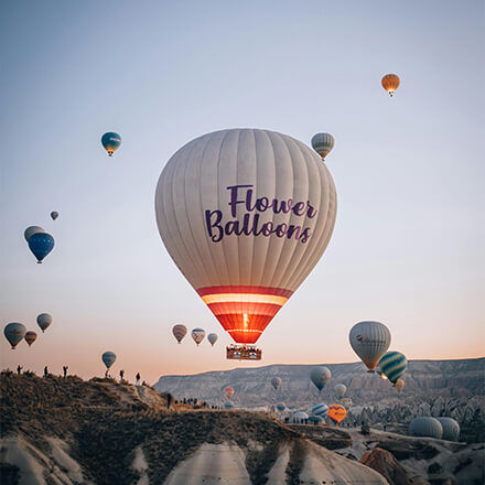 Ein großer Heißluftballon mit der Aufschrift "Flower Balloons" steigt bei Sonnenuntergang inmitten weiterer Ballons in die Luft.