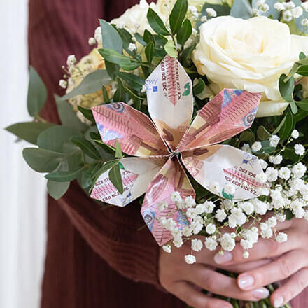 Eine gefaltete Blüte aus drei 10-Euro-Scheinen steckt in einem Blumenstrauß  von weißen Rosen, Schleierkraut und Eukalyptus