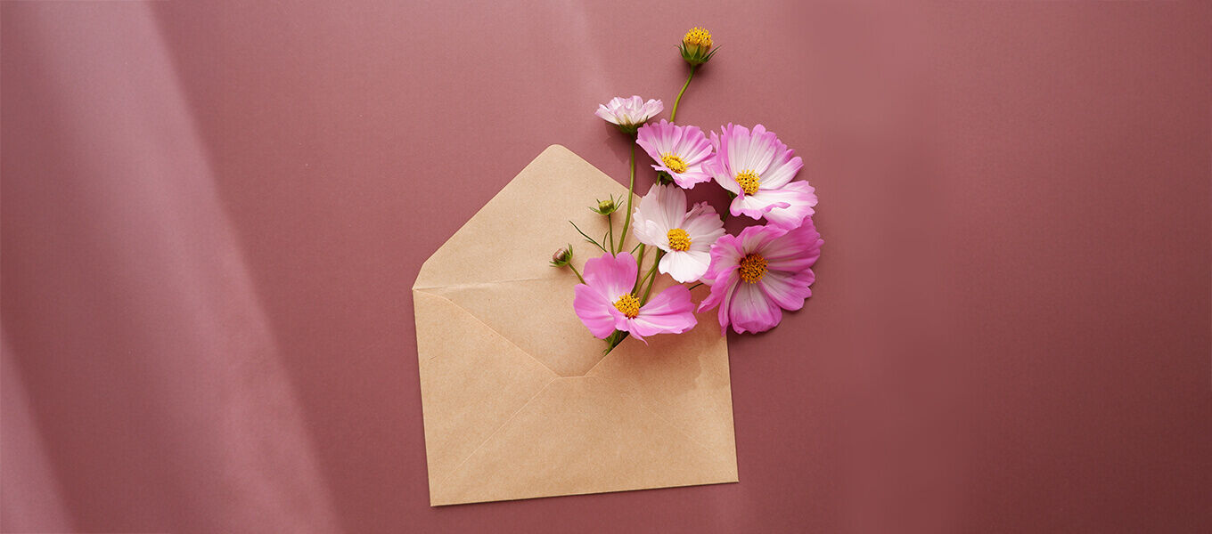 Geburtstagskarten Texte Ein brauner Umschlag liegt geöffnet auf einem rosafarbenen Hintergrund. Aus dem Umschlag ragen mehrere rosa-weiße Blüten mit gelben Staubgefäßen heraus.