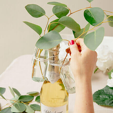 Eine Person arrangiert Eukalyptuszweige in kleinen Glasvasen, die mit Blumendraht am Hals einer Sektflasche befestigt sind. Die Sektflasche steht auf einem Tisch und ist Teil einer kreativen Hochzeitsdekoration.