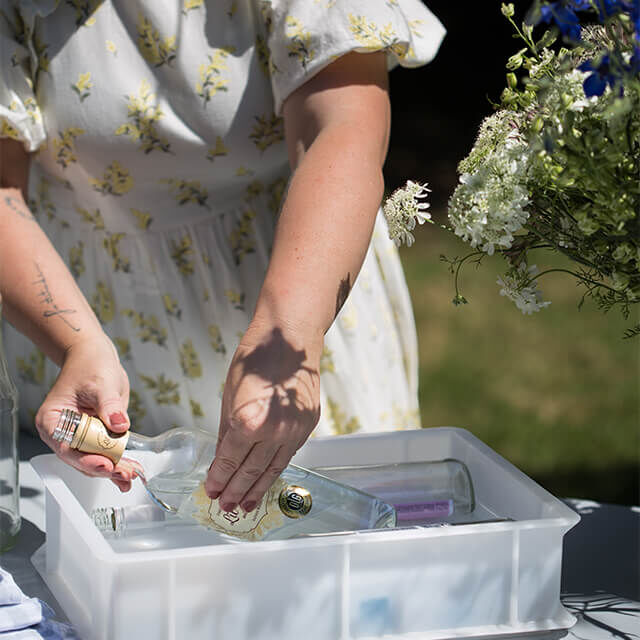 Tischdeko für Gartenparty - Step 1 Eine Frau in einem weißen Sommerkleid mit gelbem Blumenmuster taucht leere Glasflaschen in eine Plastikwanne mit Wasser.