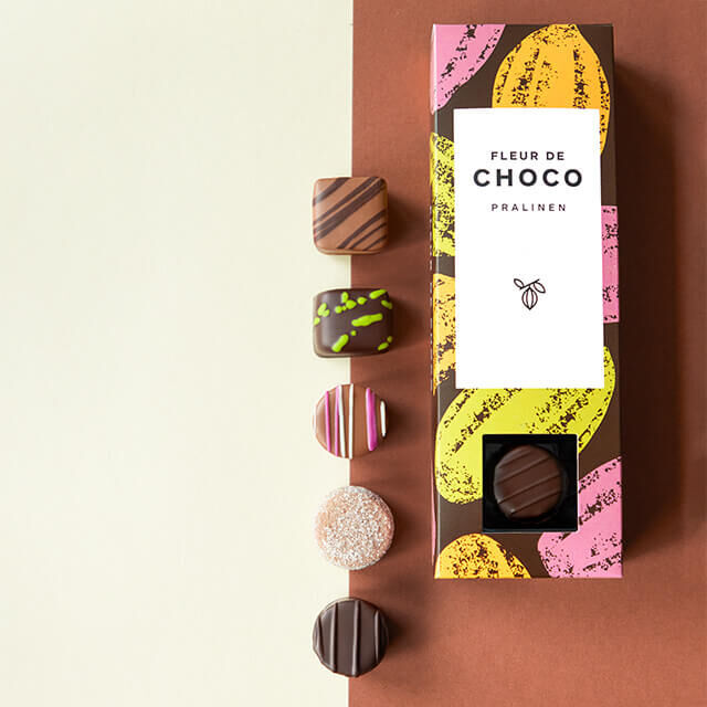 Edle Pralinen in einer stilvollen „Fleur de Choco“ Geschenkbox mit buntem Design, daneben verschiedene handverzierte Schokoladenpralinen auf braunem Hintergrund.
