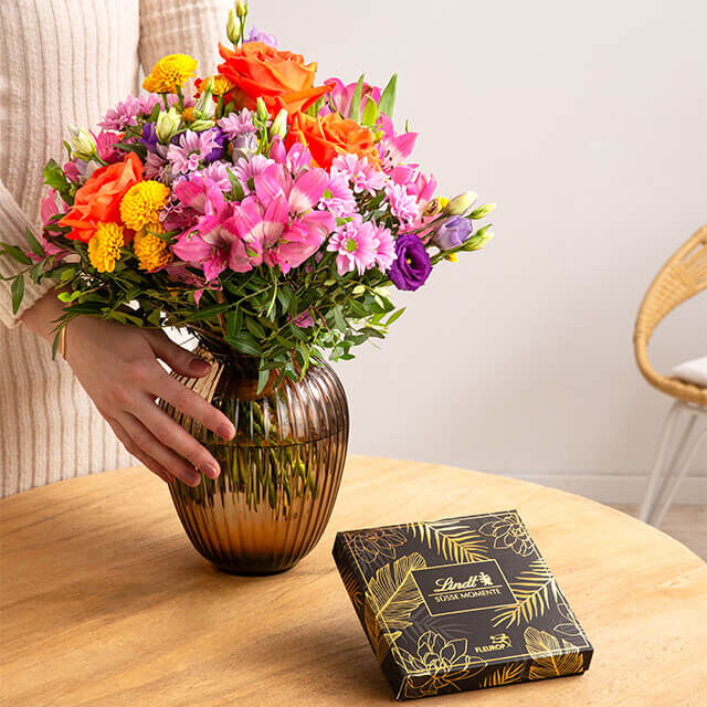 Nahaufnahme eines bunten Blumenstraußes in einer Vase auf Holztisch, daneben eine elegante Lindt-Pralinenverpackung von Fleurop