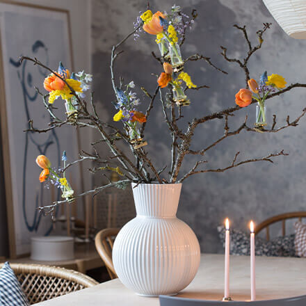 Weiße Vase gefüllt mit Ästen, an denen mithilfe kleiner Mini-Vasen Frühlingsblumen befestigt sind