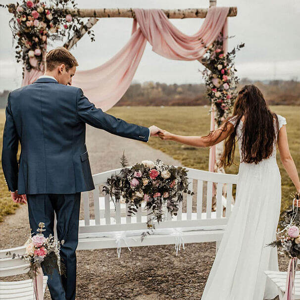 Brautpaar geht Hand in Hand durch eine Hochzeitsdekoration mit Blumenbogen bei einer freien Trauung im Freien
