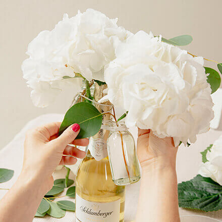 Eine Person dekoriert eine Sektflasche mit großen weißen Hortensien, die in kleinen Glasvasen arrangiert sind. Die Glasvasen sind mit Blumendraht am Flaschenhals befestigt. Diese kreative Blumendekoration dient als elegantes Hochzeitsgeschenk.