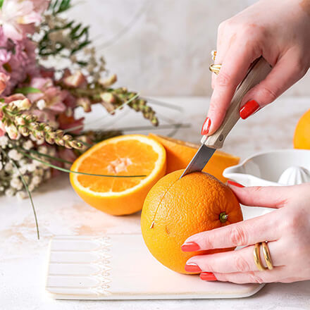 Eine Nahaufnahme einer Hand, die eine Orange auf einem weißen Schneidebrett mit einem Messer schneidet. Im Hintergrund befinden sich halbierte Orangen, eine weiße Saftpresse und unscharfe Blumen. Die Person trägt goldene Ringe und hat rote Fingernägel.