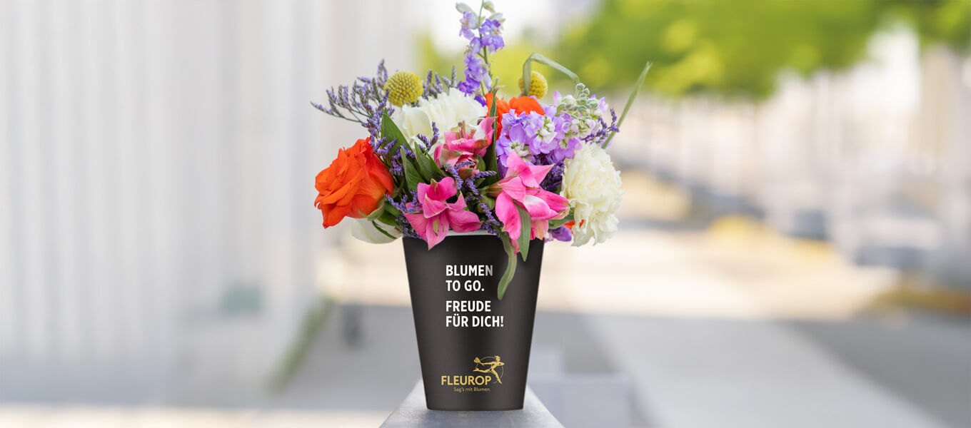 Lonely Bouquet Day Sträußchen Bunter Blumenstrauß in einem schwarzen Becher mit der Aufschrift ‚Blumen to go. Freude für Dich!‘ von Fleurop, arrangiert mit Rosen, Lilien, Nelken und Lavendel, vor urbanem, unscharfem Hintergrund.