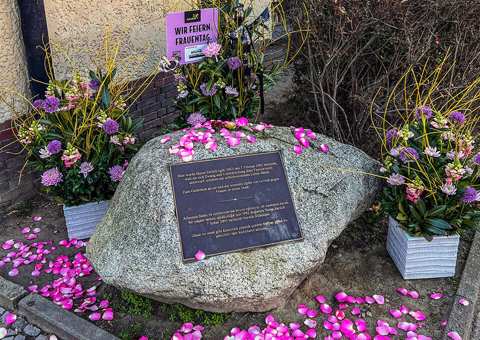 Gedenkstein für Hatun Sürücü in Berlin mit Blumen zum Frauentag Gedenkstein für Hatun Sürücü in Berlin, umrahmt von Blumengestecken in Lila und Gelb sowie Rosenblättern zum Frauentag.
