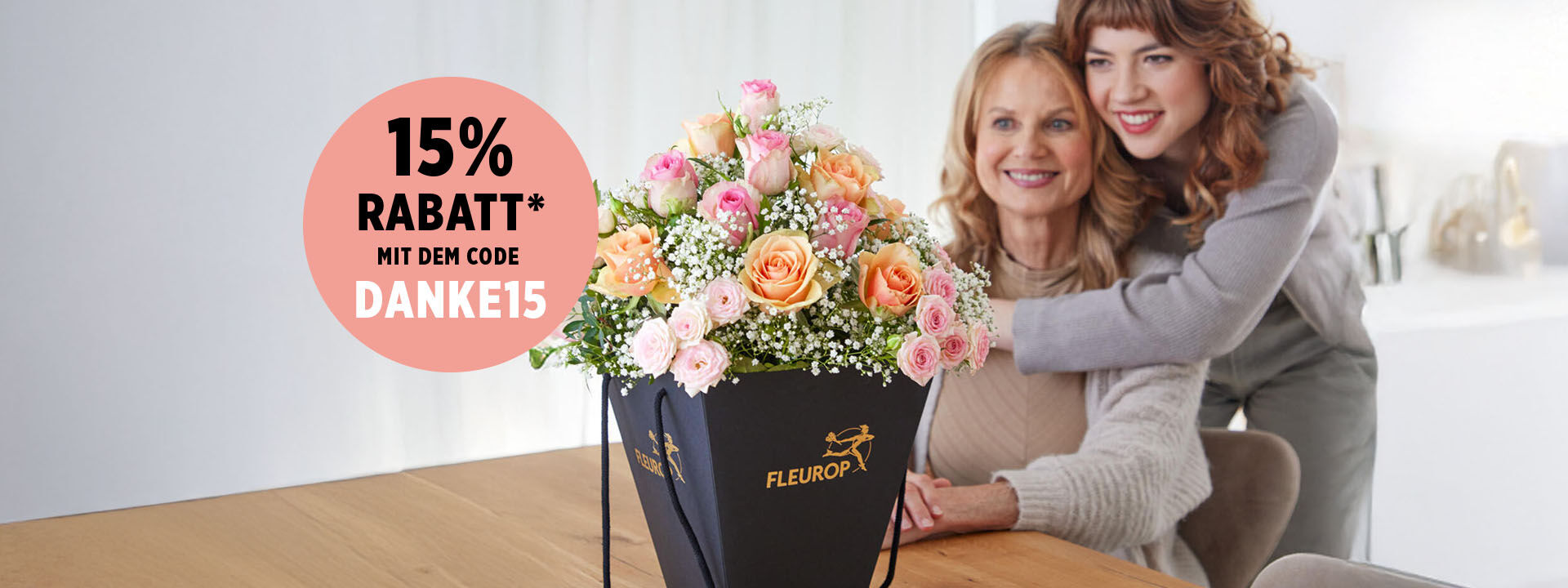 Muttertagsspecial Tochter umarmt ihre Mutter herzlich; auf dem Tisch steht ein rosa-apricotfarbener Fleurop-Blumenstrauß in einer Flowerbag – 15 % Rabatt mit dem Code DANKE15.