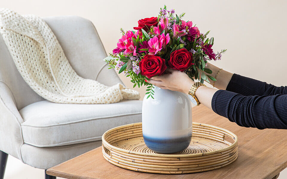 Strauß in rot-rosa mit Rosen wird in eine Vase gestellt, welche sich auf einem Wohnzimmertisch befindet.