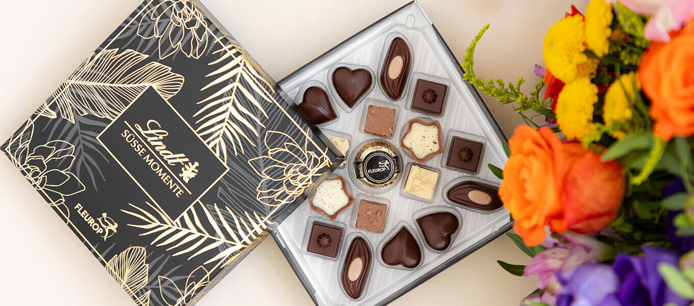 Offene Schachtel „Lindt Süße Momente“ Pralinen in edler schwarzer Verpackung mit goldenen Pflanzenmustern, daneben ein bunter Blumenstrauß mit orangefarbenen Rosen, gelben und violetten Blüten.