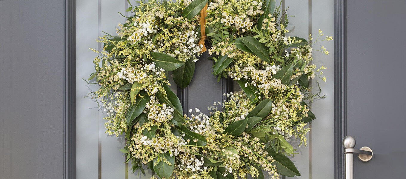 Ein wunderschöner, selbstgemachter Frühlings-Türkranz aus frischem Grün und kleinen weißen Blüten, der an einer grauen Tür hängt.