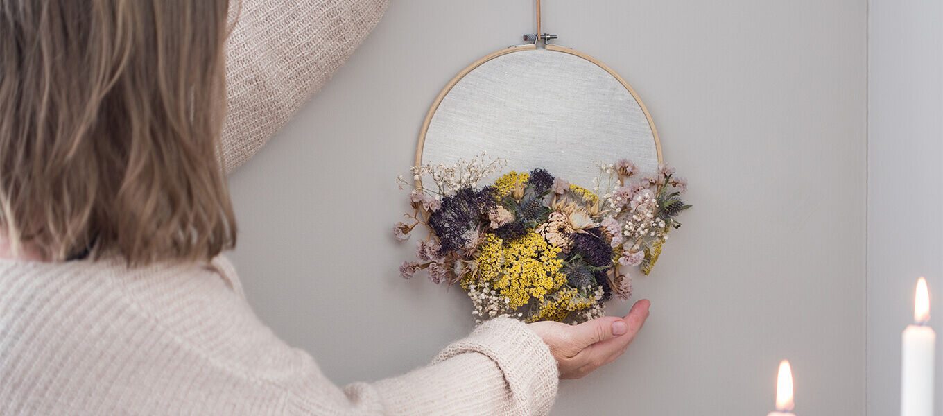 Person hängt eine dekorative Wandstickerei mit getrockneten Blumen in warmen Gelb- und Lila-Tönen auf. Im Vordergrund brennen Kerzen, was eine gemütliche Atmosphäre schafft.