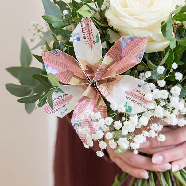 Eine gefaltete Blüte aus drei 10-Euro-Scheinen steckt in einem Blumenstrauß  von weißen Rosen, Schleierkraut und Eukalyptus