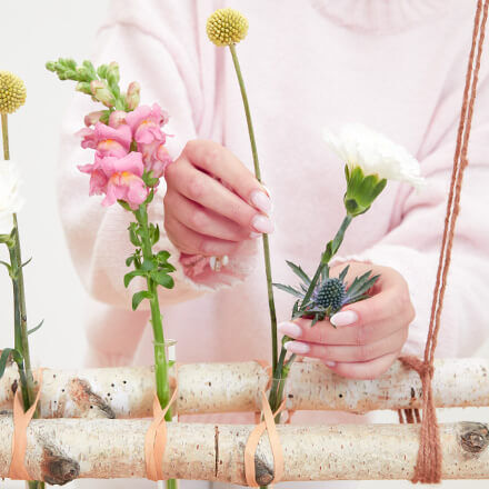 Eine Person in einem rosa Pullover arrangiert verschiedene bunte Blumen in Reagenzgläsern, die zwischen Haushaltsgummibändern an zwei Birkenstämmen befestigt sind.