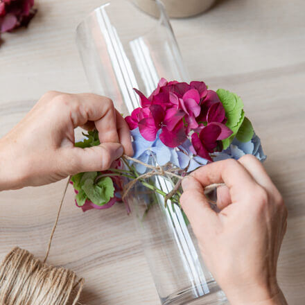 Zwei Hände machen einen Knoten um ein Zylinderglasgefäß. Zuvor wurden mit der Juteschnur kleine Teile der Hortensienblüte am Glas befestigt.