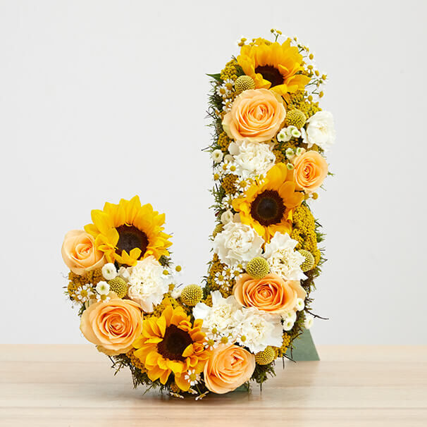 Ein dekorativer Buchstabe "J" aus frischen Blumen, darunter Sonnenblumen, Rosen und andere Blüten, auf einem Holztisch. Die Blumen sind dicht aneinander gereiht und bilden eine lebendige und farbenfrohe Gestaltung.