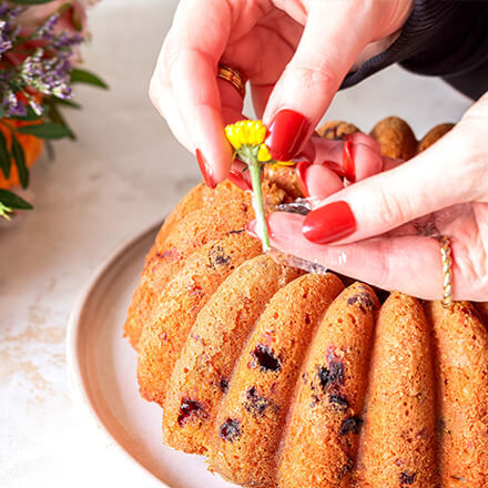 Ein frisch gebackener Gugelhupf auf einem Teller, der mit kleinen Blüten dekoriert wird. Eine Person mit roten Fingernägeln steckt eine gelbe Blume vorsichtig auf den Kuchen. Im Hintergrund sind orangefarbene Blumen und grüne Blätter zu sehen.