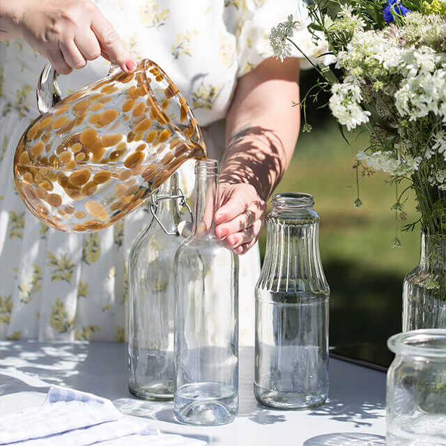 Tischdeko für Gartenparty - Step 3 Eine Frau gießt mit einem gemusterten Glaskrug Wasser in eine der vorbereiteten Glasflaschen.