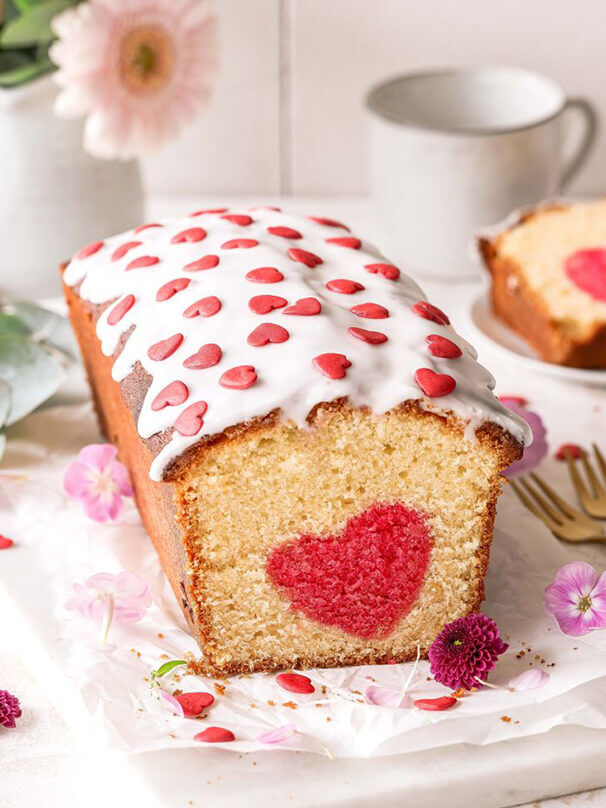 Aufgeschnittener Kuchen mit Herzüberraschung im Inneren, dekoriert mit Zuckerguss und Zuckerherzen, daneben Blüten und Besteck.