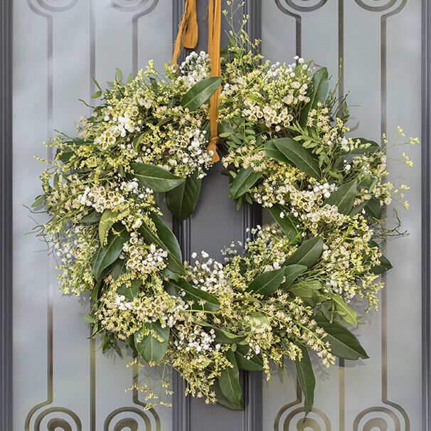 Ein wunderschöner, selbstgemachter Frühlings-Türkranz aus frischem Grün und kleinen weißen Blüten, der an einer grauen Tür hängt.