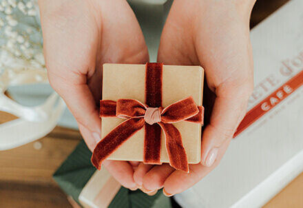 Hände halten ein kleines Geschenk in braunem Papier mit roter Schleife.