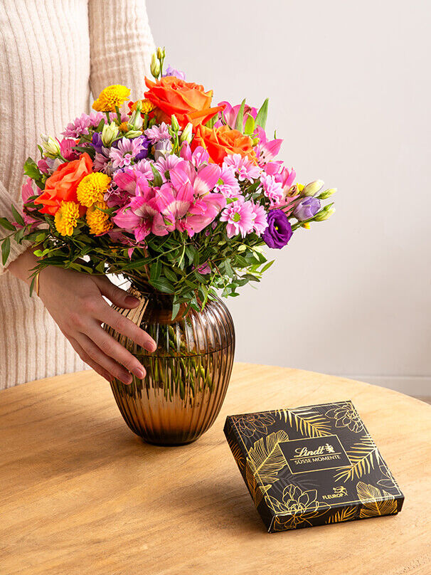 Nahaufnahme eines bunten Blumenstraußes in einer Vase auf Holztisch, daneben eine elegante Lindt-Pralinenverpackung von Fleurop