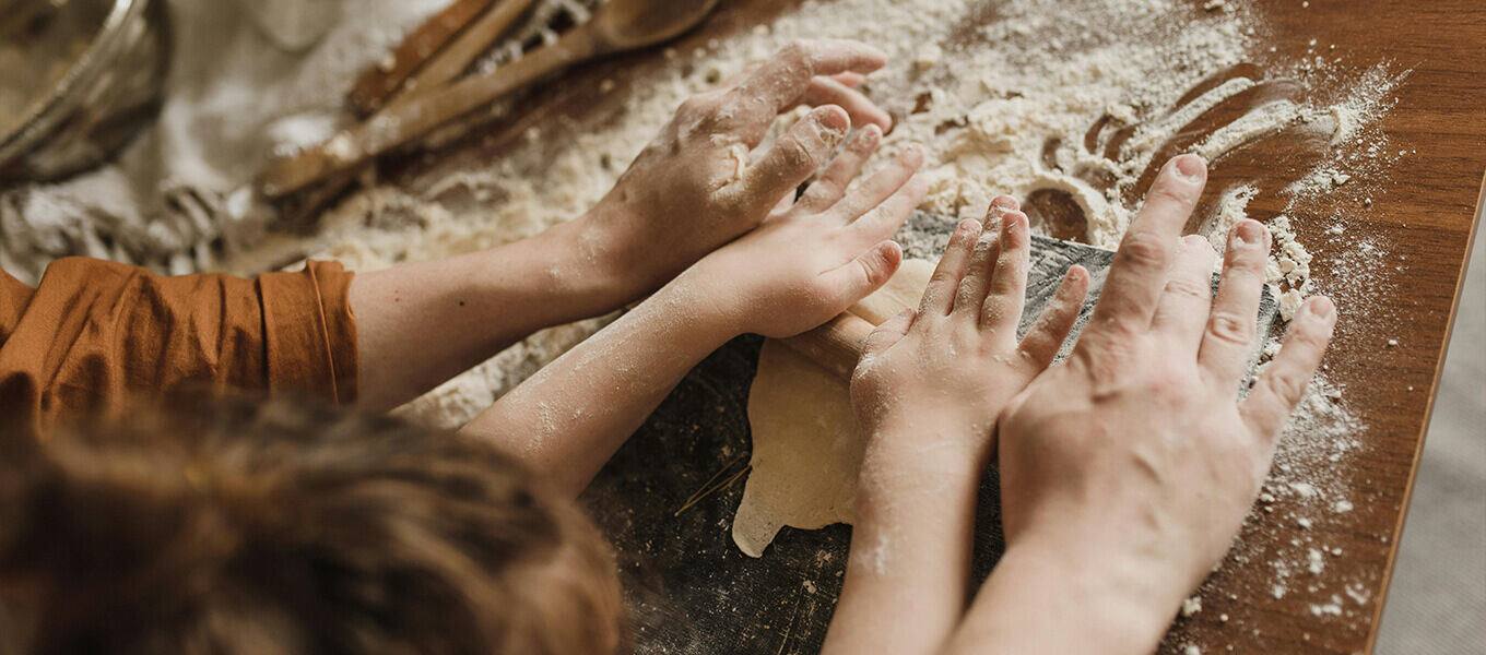 Mehrere Hände – von Erwachsenen und einem Kind – rollen gemeinsam Teig auf einer bemehlten Arbeitsfläche aus. Die Szene vermittelt eine warme, familiäre Atmosphäre beim gemeinsamen Backen. Im Hintergrund liegen Holzlöffel und Backutensilien.