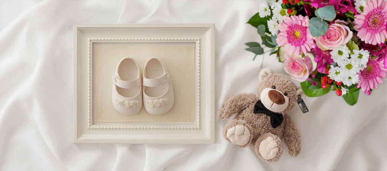 Blumenstrauß zur Geburt mit rosa Gerbera, Rosen und Teddybär neben Babyschuhen