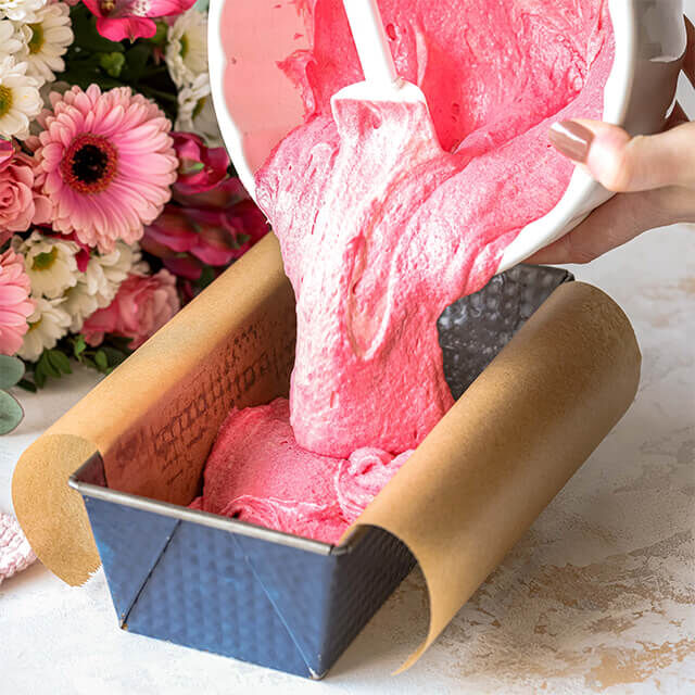 Herzkuchen Step 1 Rosa Kuchenteig wird in eine mit Backpapier ausgelegte Kastenform gefüllt, im Hintergrund ein Strauß aus pink-weißen Blumen.