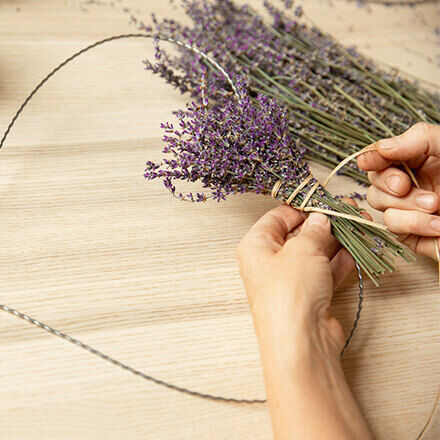 Eine Person befestigt einen Strauß Lavendel mit einem Band an einem herzförmigen Drahtrahmen. Weitere Lavendelblüten liegen auf dem Tisch, während der Lavendel schrittweise um den Rahmen gebunden wird.