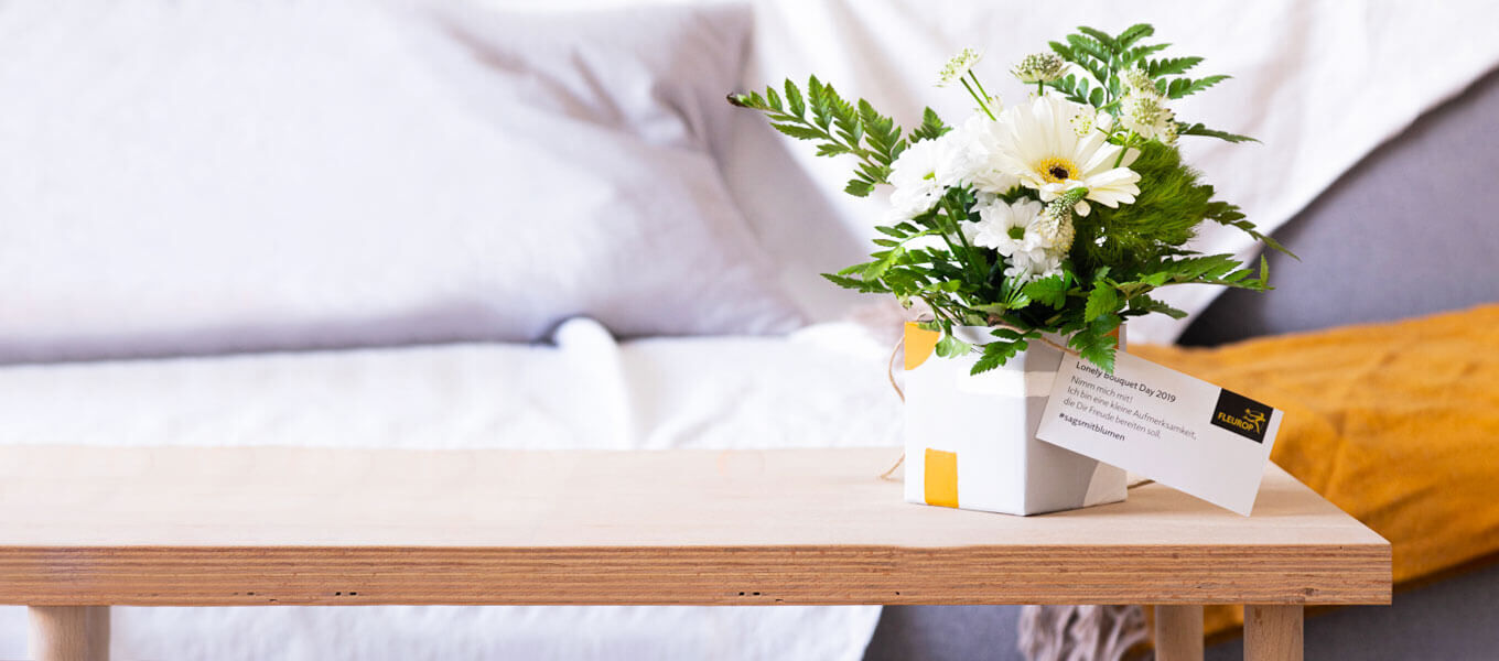 Ein Blumensträußchen in Weiß steht in einem schön bemalten Getränkekarton auf einem Tischchen vor dem Sofa