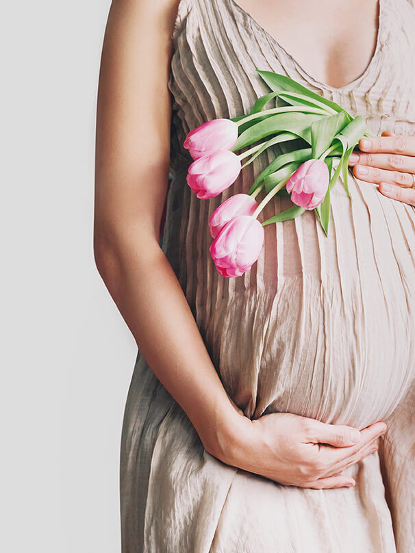 Schwangere Frau im hellen Kleid hält rosa Tulpen vor ihrem Babybauch als Symbol für Glückwünsche zur Geburt und neues Leben