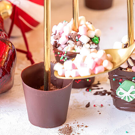 Schokoladen-Cups, die mit bunten Streuseln, Mini-Marshmallows und kleinen Weihnachtsdekorationen verziert werden.