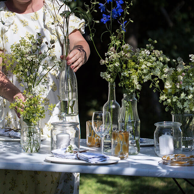 Tischdeko für Gartenparty - Step 5 Sie arrangiert verschiedene Wildblumen in Flaschen und Gläsern auf einem gedeckten Tisch im Garten.