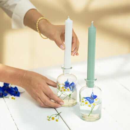 Eine weiße Kerze wird auf ein Ginglas gesteckt, welches mit etwas Wasser, Kamille- und Ritterspornblüten gefüllt ist. Daneben steht eine weitere Kerze mit einem Ginglas. Diese Kerze ist in einem Min Ton.