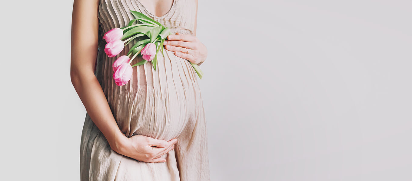 Schwangere Frau mit rosa Tulpen vor dem Babybauch als emotionales Headerbild für Glückwünsche zur Geburt