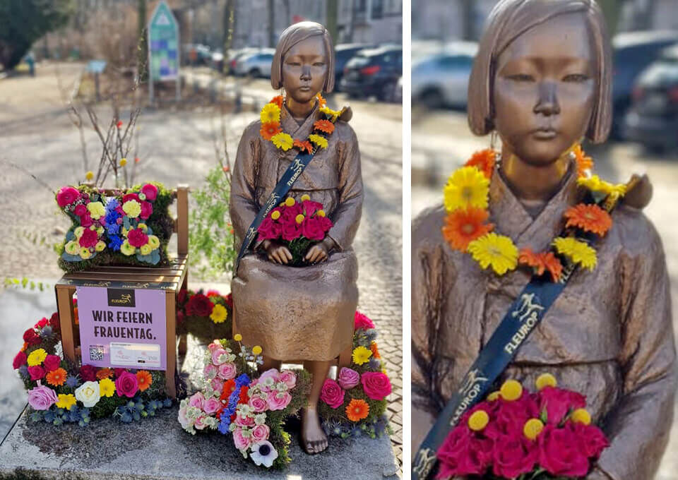 Statue eines Mädchens auf einem Stuhl in Berlin, umgeben von bunten Blumen und Blütenketten, dekoriert zum Internationalen Frauentag.