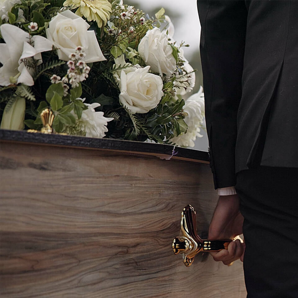 Sarg mit weißem Trauerblumenarrangement aus Rosen und Lilien, getragen von einem schwarz gekleideten Träger